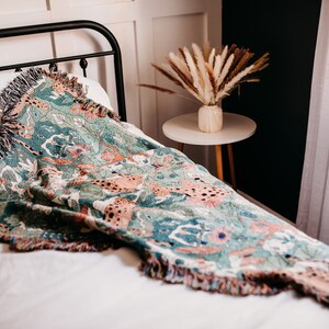 May include: A green, pink, and brown woven throw blanket with a fringe border. The blanket is draped over a white bed with a black metal headboard. A vase with dried pampas grass sits on a small round table next to the bed.