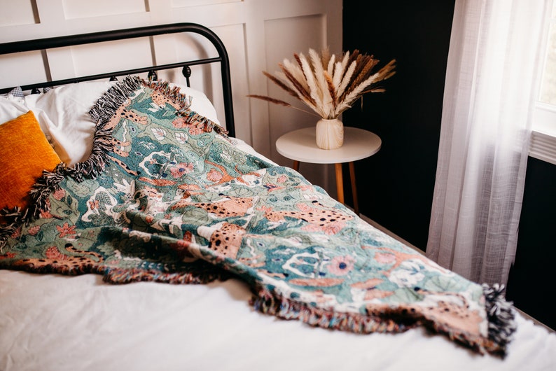 May include: A green, pink, and brown woven throw blanket with a fringe border. The blanket is draped over a white bed with a black metal headboard. A vase with dried pampas grass sits on a small round table next to the bed.
