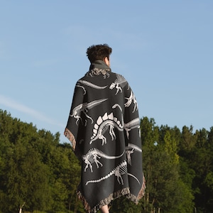 May include: A person wearing a black and white dinosaur skeleton patterned throw blanket walks on a grassy field.