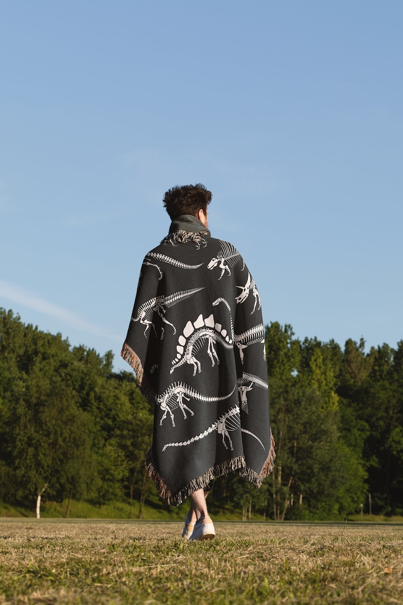 May include: A person wearing a black and white dinosaur skeleton patterned throw blanket walks on a grassy field.