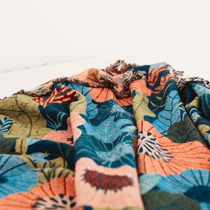 May include: A colorful woven throw blanket with a floral pattern in shades of blue, green, orange, and pink. The blanket has a fringed edge.