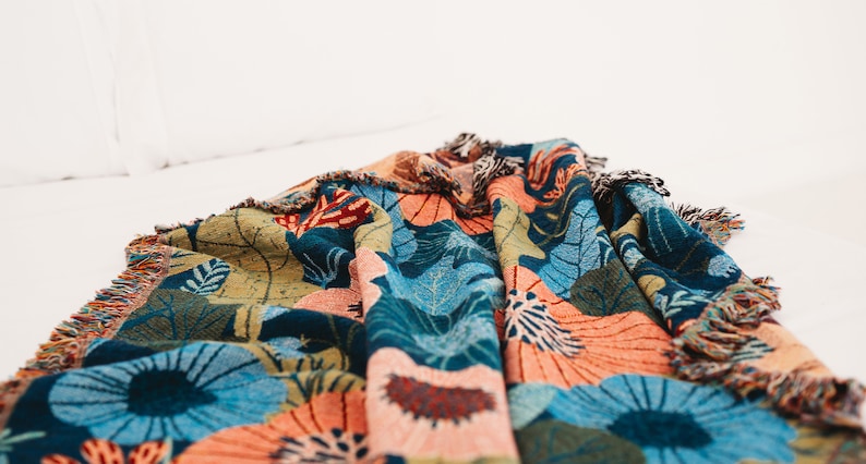 May include: A colorful woven throw blanket with a floral pattern in shades of blue, green, orange, and pink. The blanket has a fringed edge.
