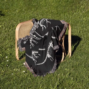 May include: A black throw blanket with a white dinosaur skeleton pattern draped over a wooden chair. The chair is sitting on a green grassy lawn.