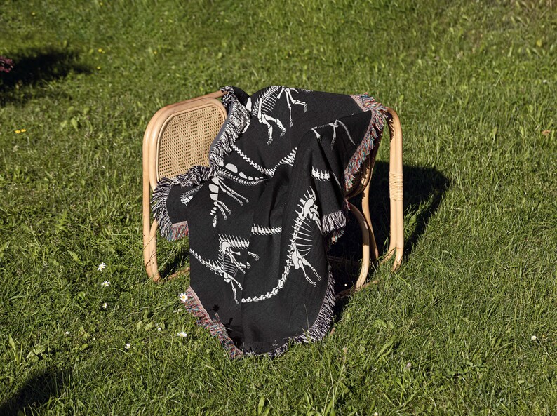 May include: A black throw blanket with a white dinosaur skeleton pattern draped over a wooden chair. The chair is sitting on a green grassy lawn.