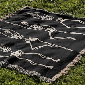 May include: Black throw blanket with a white skeleton pattern. The skeletons are dancing and have skulls. The blanket has a fringe border.