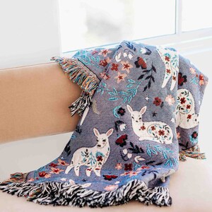 May include: A soft, blue throw blanket with a woven design of white deer and floral patterns. The blanket has fringe on all sides.