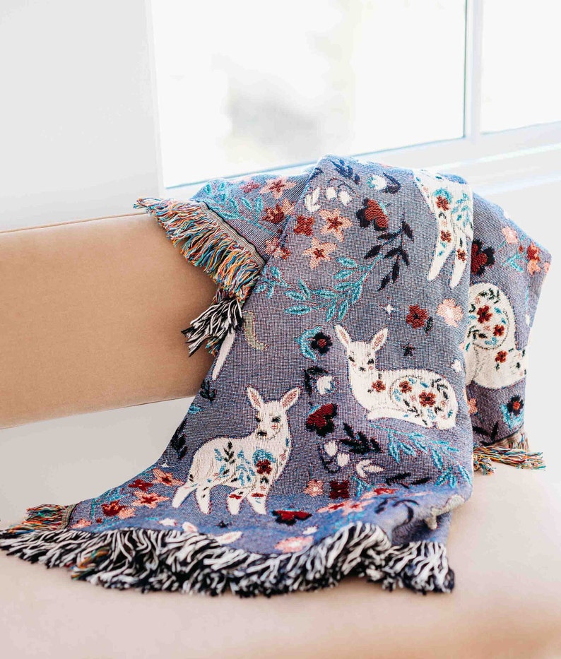 May include: A soft, blue throw blanket with a woven design of white deer and floral patterns. The blanket has fringe on all sides.