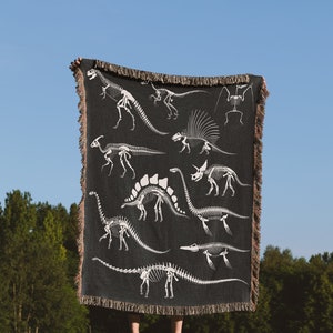 May include: A black woven throw blanket with a white dinosaur skeleton pattern. The blanket is being held up by a person in a grassy field.