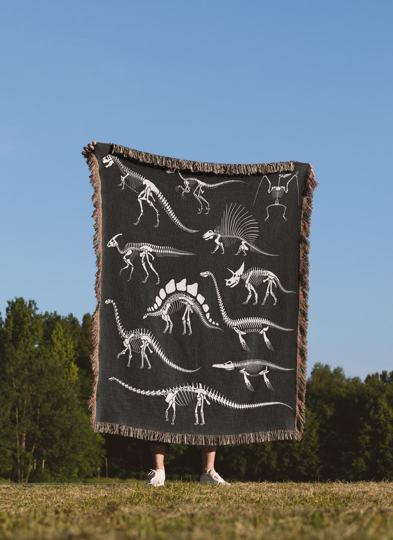 May include: A black woven throw blanket with a white dinosaur skeleton pattern. The blanket is being held up by a person in a grassy field.