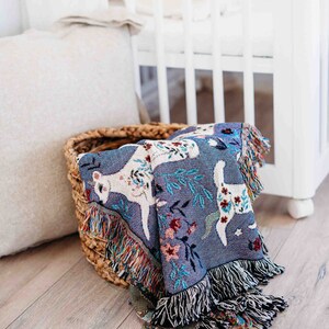 May include: A woven throw blanket with a blue background and a white deer pattern. The blanket is folded and placed in a wicker basket.