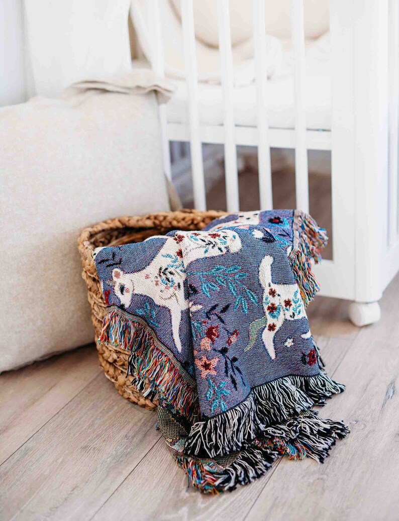 May include: A woven throw blanket with a blue background and a white deer pattern. The blanket is folded and placed in a wicker basket.