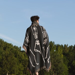 May include: A person wearing a black and white skeleton patterned throw blanket walks on a grassy field. The blanket has fringe around the edges.
