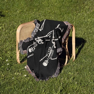May include: A black woven throw blanket with a white skeleton design. The blanket is draped over a wooden chair with a woven back.