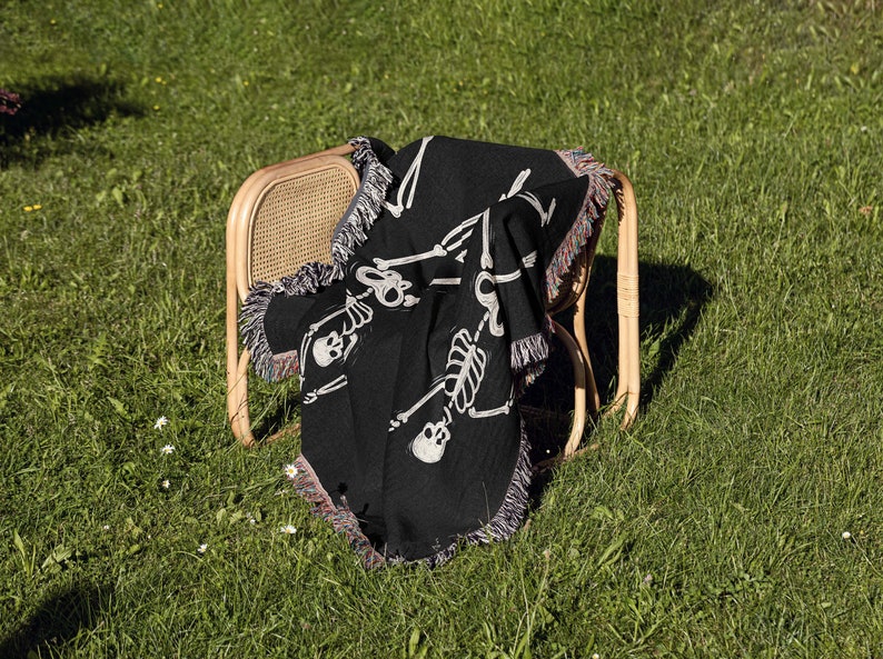 May include: A black woven throw blanket with a white skeleton design. The blanket is draped over a wooden chair with a woven back.