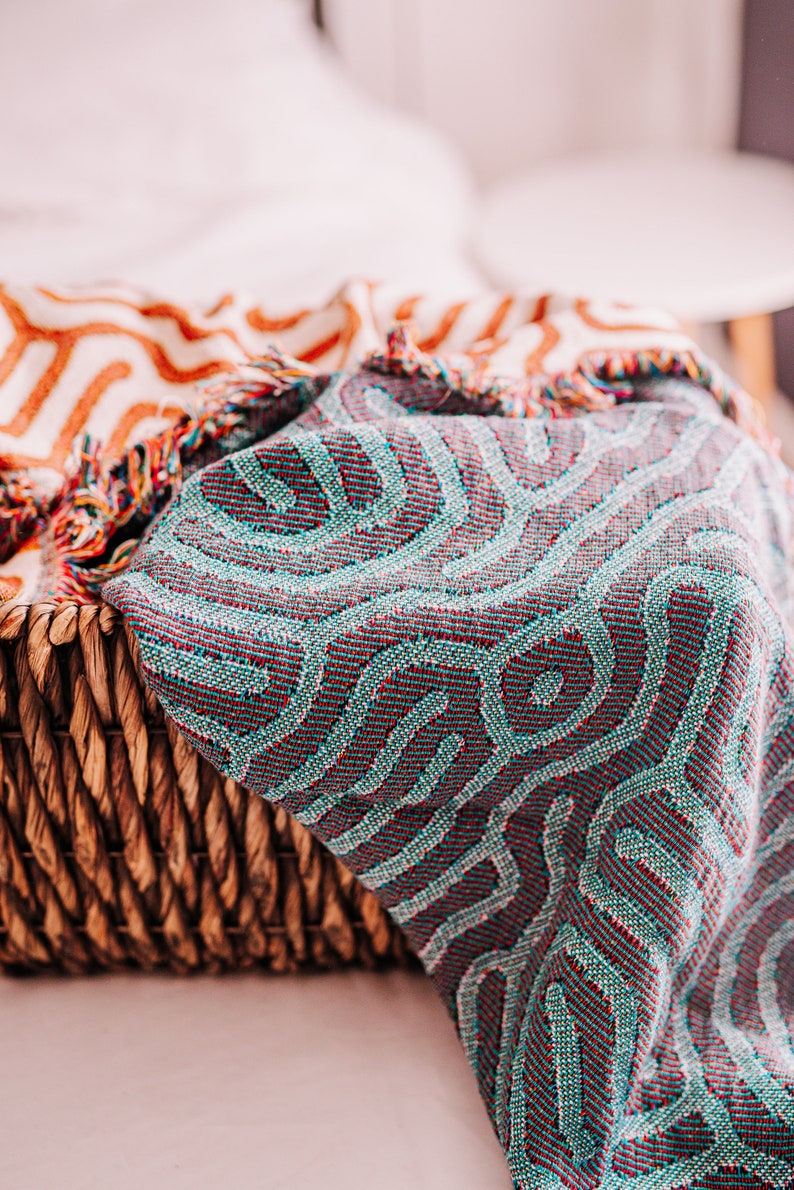 Peut inclure: Une couverture en tissu tiss&eacute; avec un motif g&eacute;om&eacute;trique bleu sarcelle et rouge. La couverture est pos&eacute;e sur un panier en osier.