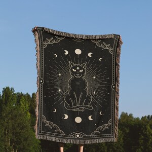 May include: A black woven tapestry with a gold design of a cat with three eyes sitting in front of a crescent moon and stars. The tapestry is held up by a person in front of a blue sky and green trees.