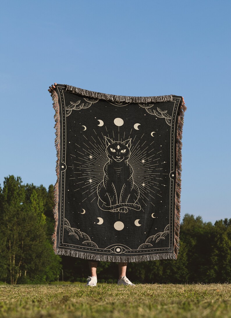 May include: A black woven tapestry with a gold design of a cat with three eyes sitting in front of a crescent moon and stars. The tapestry is held up by a person in front of a blue sky and green trees.