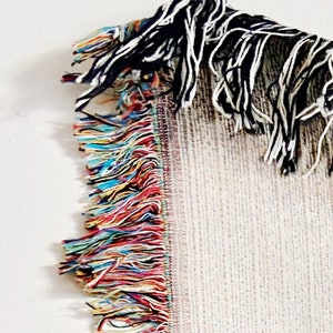 Puede incluir: Primer plano del fleco de una manta tejida. El fleco es una mezcla de hilos de colores, incluyendo rojo, azul, amarillo y verde. La manta es blanca con un patr&oacute;n sutil.