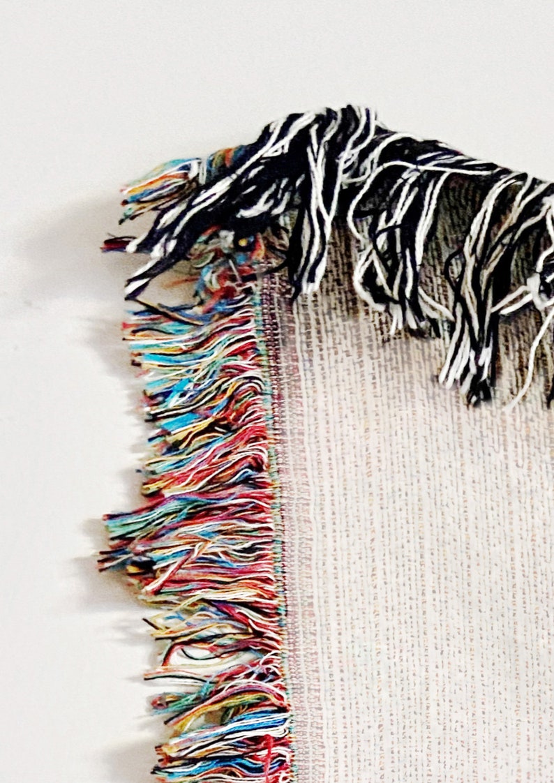Puede incluir: Primer plano del fleco de una manta tejida. El fleco es una mezcla de hilos de colores, incluyendo rojo, azul, amarillo y verde. La manta es blanca con un patr&oacute;n sutil.