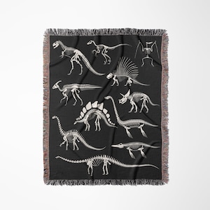 May include: A black woven throw blanket with a white silhouette design of various dinosaur skeletons. The skeletons are arranged in a random pattern on the blanket.