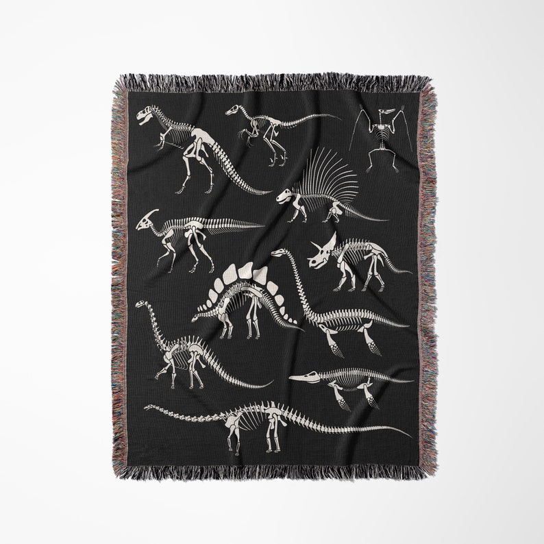 May include: A black woven throw blanket with a white silhouette design of various dinosaur skeletons. The skeletons are arranged in a random pattern on the blanket.