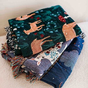 May include: A stack of three woven throw blankets with fringe. The top blanket is green with a deer and floral pattern. The middle blanket is blue with a white deer and floral pattern. The bottom blanket is dark blue with a white star pattern.