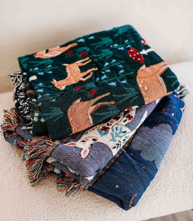 May include: A stack of three woven throw blankets with fringe. The top blanket is green with a deer and floral pattern. The middle blanket is blue with a white deer and floral pattern. The bottom blanket is dark blue with a white star pattern.