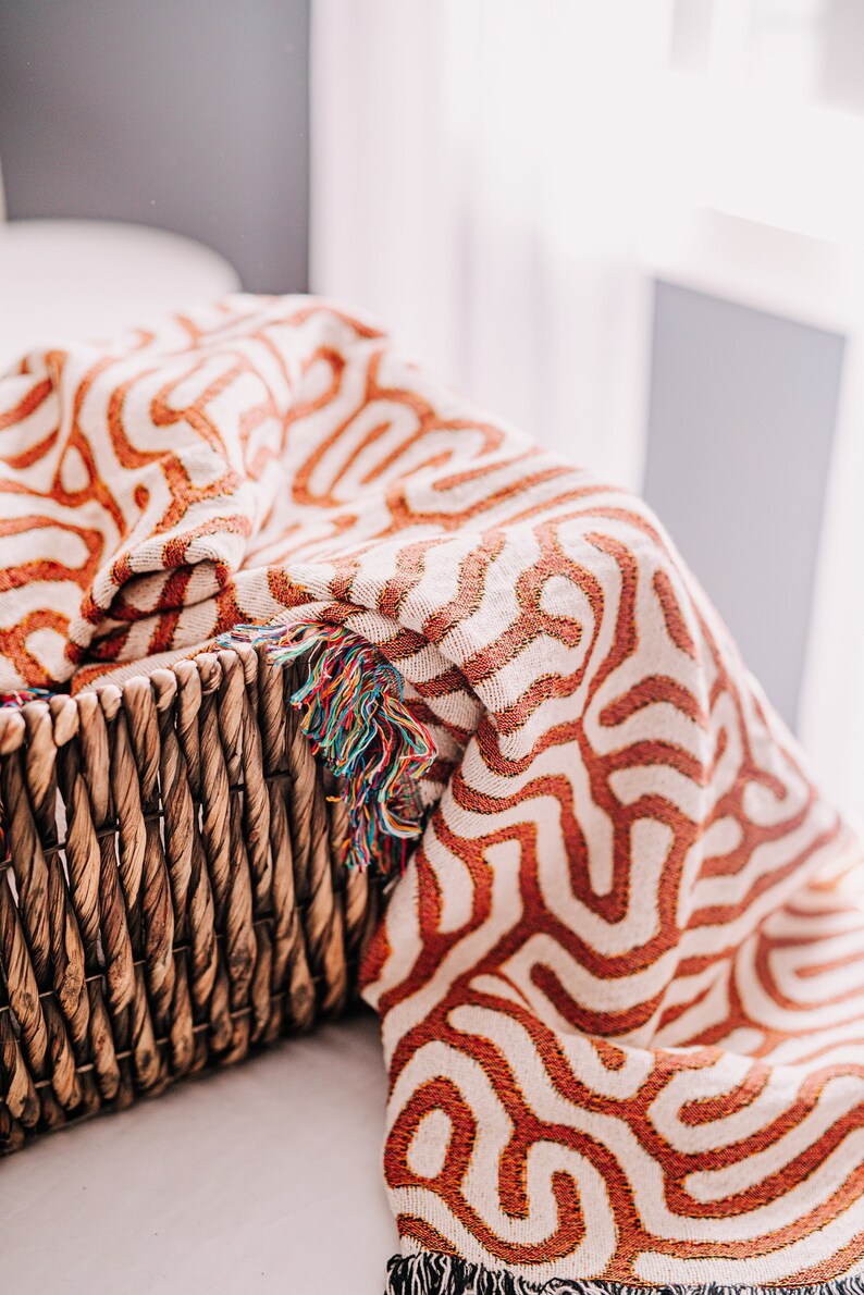 Peut inclure: Une couverture en laine tiss&eacute;e avec un motif g&eacute;om&eacute;trique dans les tons orange et blanc. La couverture a une bordure en franges et est drap&eacute; sur un panier en osier.