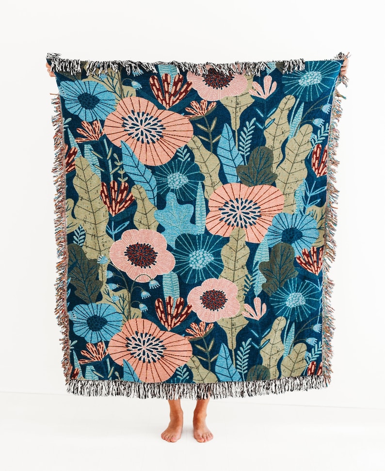 May include: A woven throw blanket with a floral pattern in shades of pink, blue, and green on a dark blue background. The blanket has fringe on all sides.