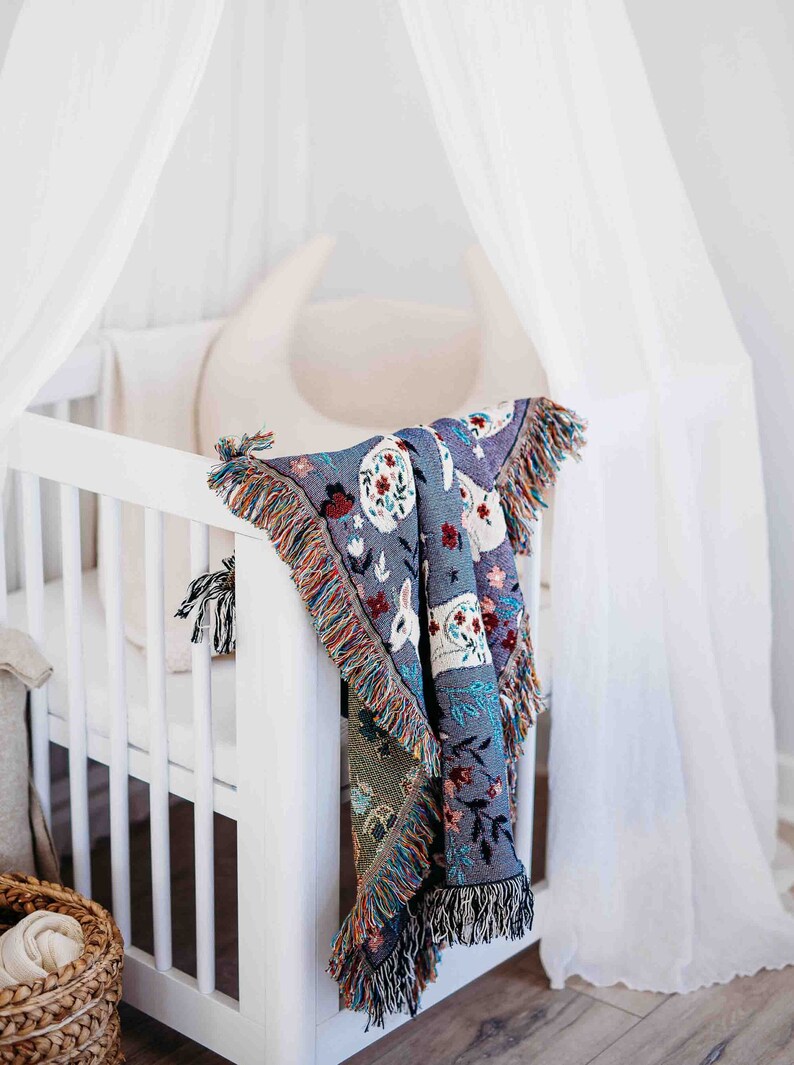 May include: A white crib with a white canopy and a colorful woven blanket hanging over the side. The blanket has a floral and animal design.