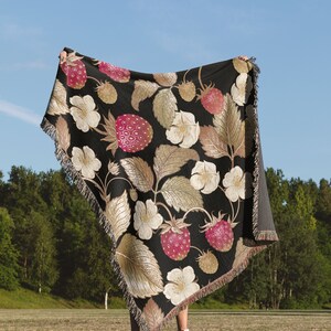 May include: A black throw blanket with a strawberry pattern. The blanket features large pink strawberries, white flowers, and gold-toned leaves. The blanket is fringed and held up against a blue sky and green background.