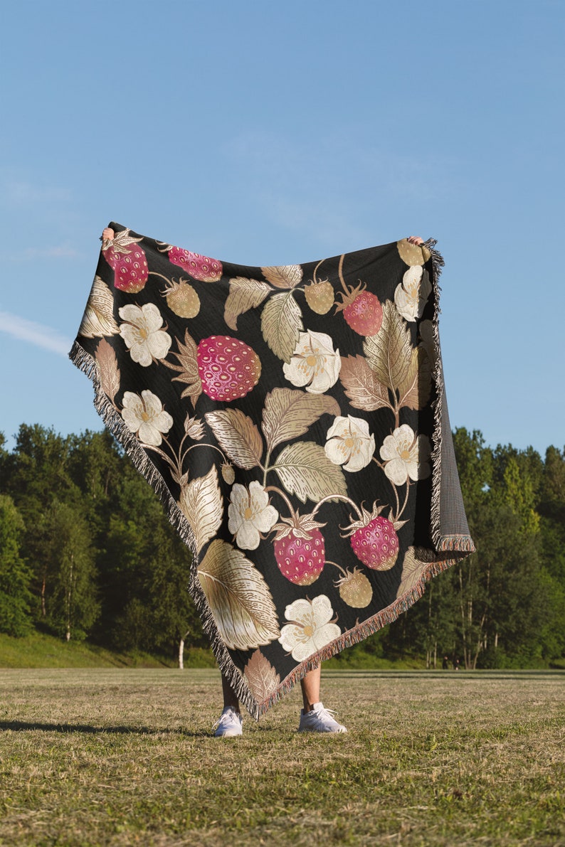 May include: A black throw blanket with a strawberry pattern. The blanket features large pink strawberries, white flowers, and gold-toned leaves. The blanket is fringed and held up against a blue sky and green background.