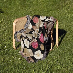 May include: A black throw blanket draped over a wicker chair, featuring a vibrant strawberry and floral pattern. The blanket has a fringe trim and is set against a backdrop of green grass, creating a cozy, summery scene.