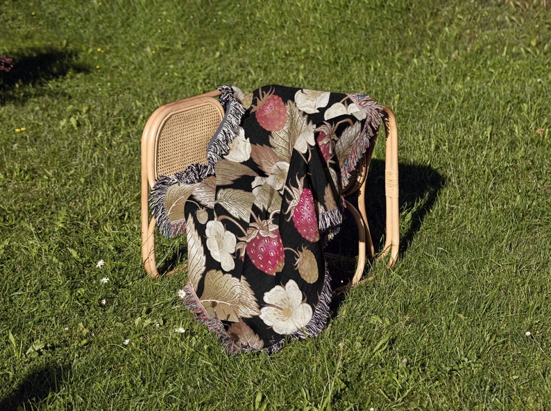 May include: A black throw blanket draped over a wicker chair, featuring a vibrant strawberry and floral pattern. The blanket has a fringe trim and is set against a backdrop of green grass, creating a cozy, summery scene.