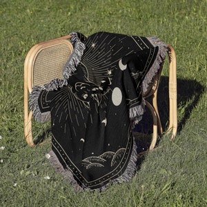 May include: Black woven throw blanket with a celestial design featuring a hand reaching towards a crescent moon and stars. The blanket has fringe on all sides and is draped over a wicker chair.