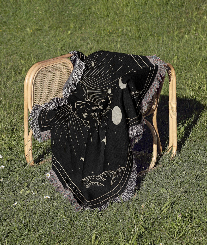May include: Black woven throw blanket with a celestial design featuring a hand reaching towards a crescent moon and stars. The blanket has fringe on all sides and is draped over a wicker chair.