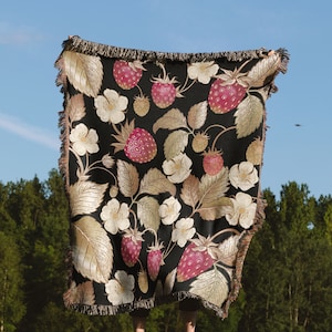 May include: A black throw blanket with a repeating pattern of strawberries, leaves, and white flowers. The strawberries are red and the leaves are gold. The blanket has a fringed edge and is held up against a blue sky.