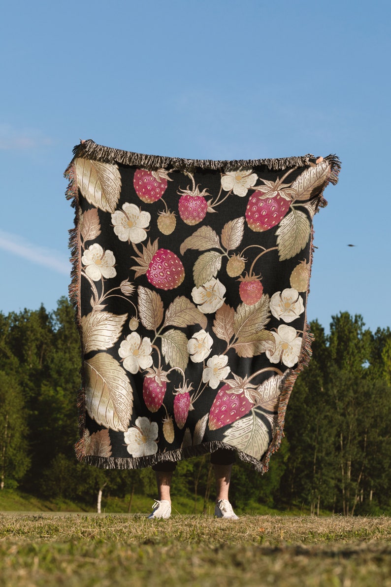 May include: A black throw blanket with a repeating pattern of strawberries, leaves, and white flowers. The strawberries are red and the leaves are gold. The blanket has a fringed edge and is held up against a blue sky.