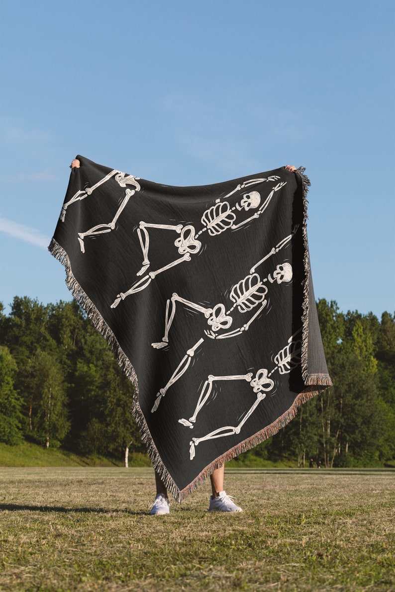 May include: A black woven throw blanket with a white skeleton design. The skeleton is in a dancing pose. The blanket is being held up by a person in a grassy field.