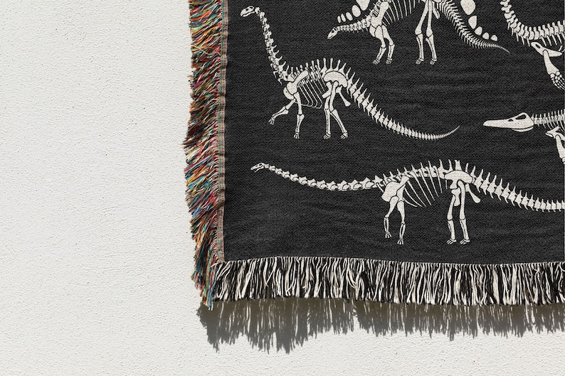 May include: Black woven throw blanket with a white dinosaur skeleton pattern. The blanket has a fringe border.