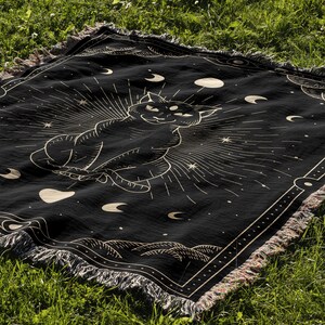 May include: Black woven throw blanket with a gold outline of a cat sitting in a circle of moons and stars. The blanket has a fringe border.
