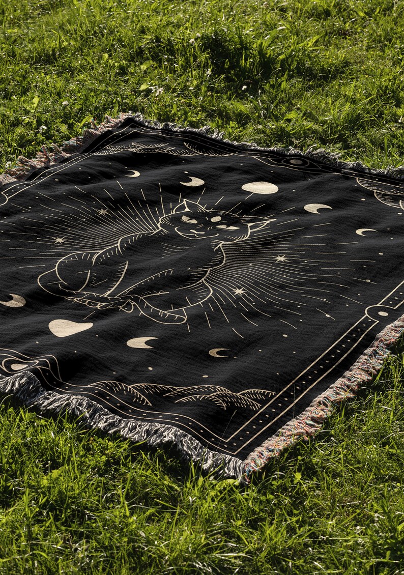 May include: Black woven throw blanket with a gold outline of a cat sitting in a circle of moons and stars. The blanket has a fringe border.