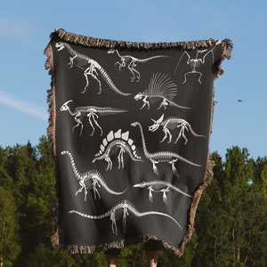 May include: A person holding a black woven throw blanket with a white dinosaur skeleton pattern. The blanket has fringe on the edges.