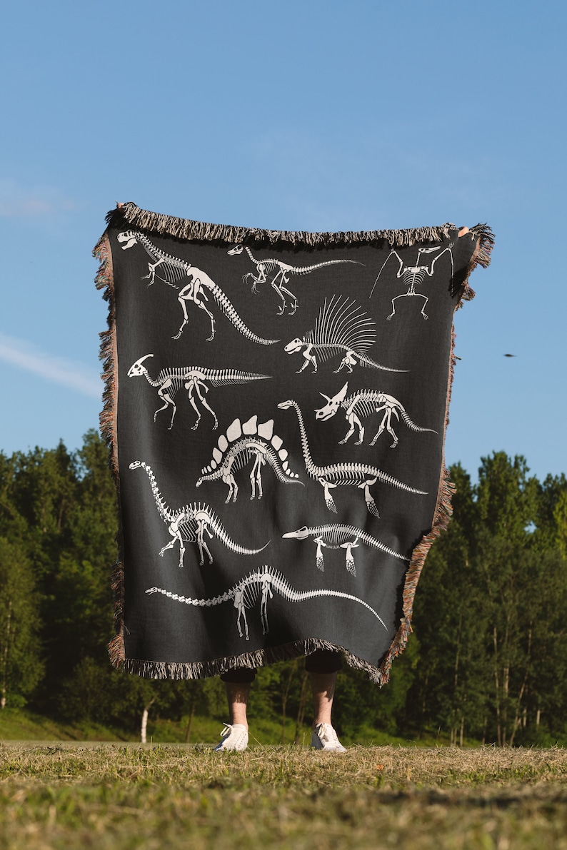 May include: A person holding a black woven throw blanket with a white dinosaur skeleton pattern. The blanket has fringe on the edges.