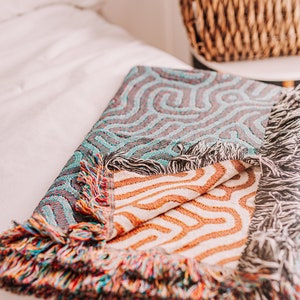 Peut inclure: Une couverture en tissage color&eacute;e avec un motif g&eacute;om&eacute;trique dans les tons de bleu, orange et blanc. La couverture a une bordure &agrave; franges et est pli&eacute;e sur un lit blanc.