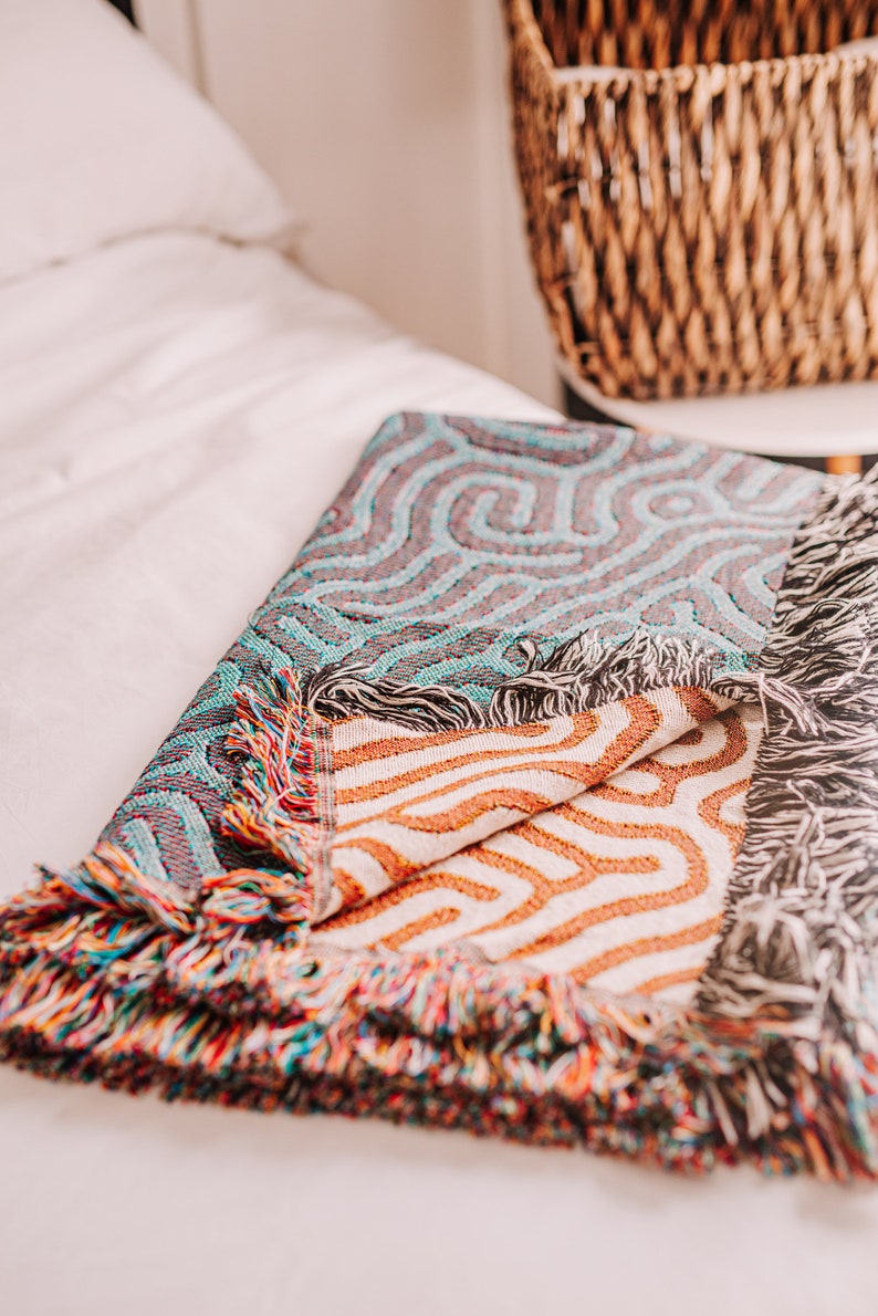 Peut inclure: Une couverture en tissage color&eacute;e avec un motif g&eacute;om&eacute;trique dans les tons de bleu, orange et blanc. La couverture a une bordure &agrave; franges et est pli&eacute;e sur un lit blanc.
