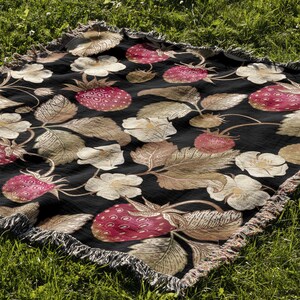 May include: A black woven blanket with a repeating pattern of strawberries, leaves, and flowers in shades of red, beige, and white. The blanket has a fringed edge and is displayed on green grass.