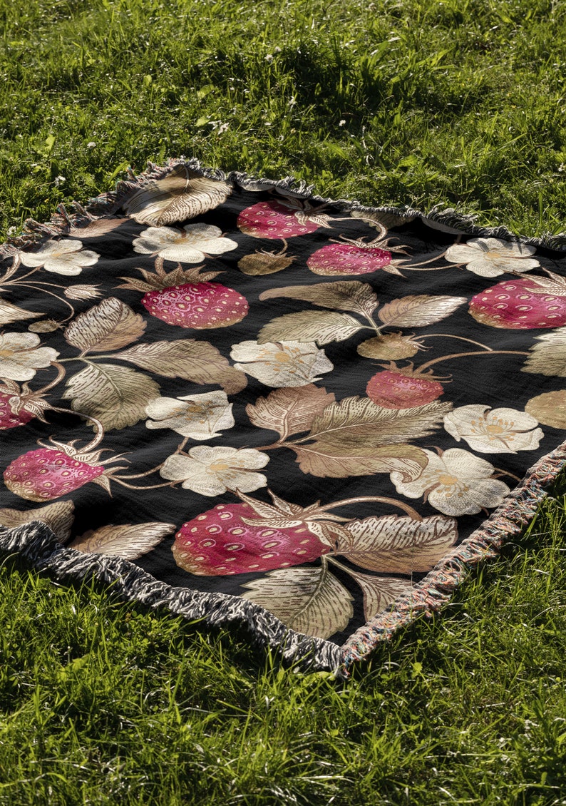 May include: A black woven blanket with a repeating pattern of strawberries, leaves, and flowers in shades of red, beige, and white. The blanket has a fringed edge and is displayed on green grass.