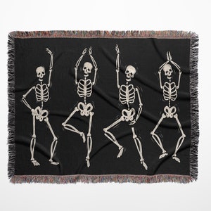 May include: A black woven throw blanket with a design of four white skeletons dancing. The skeletons are in various poses with their arms raised.