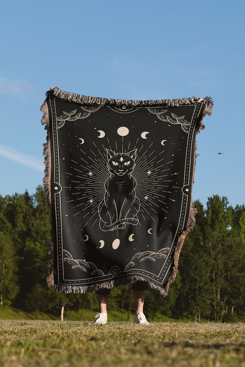 May include: A black woven tapestry with a gold design of a cat with three eyes, surrounded by moons and stars. The tapestry is being held up by a person in front of a green grassy field and trees.